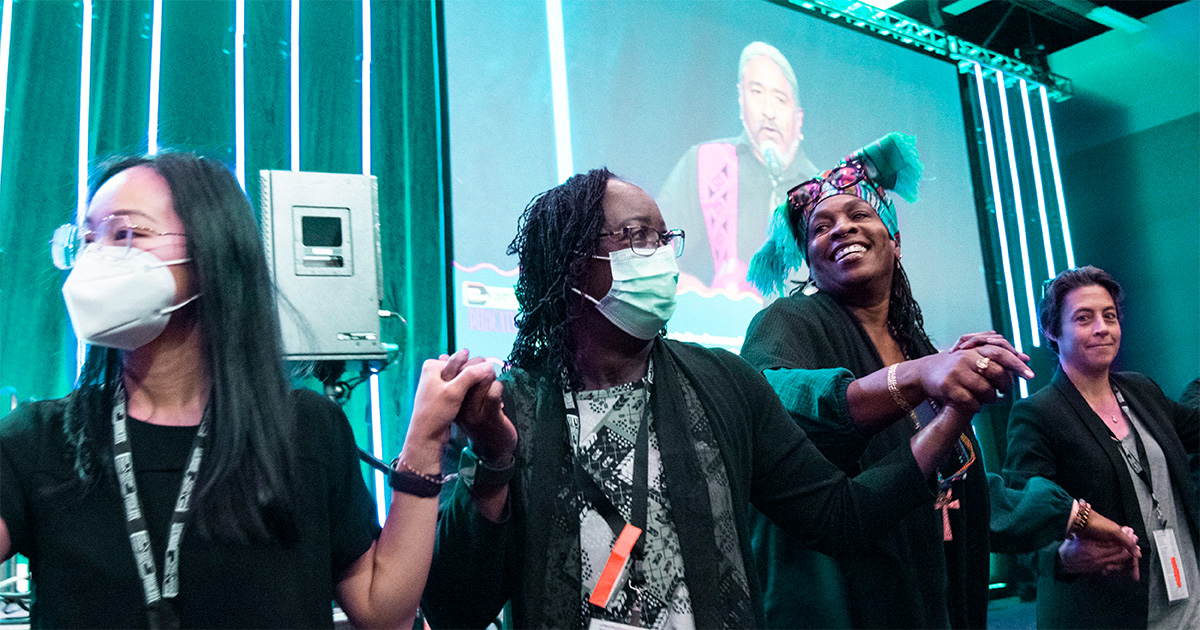 Four people holding hands in front of a stage with speaker on screen in background.