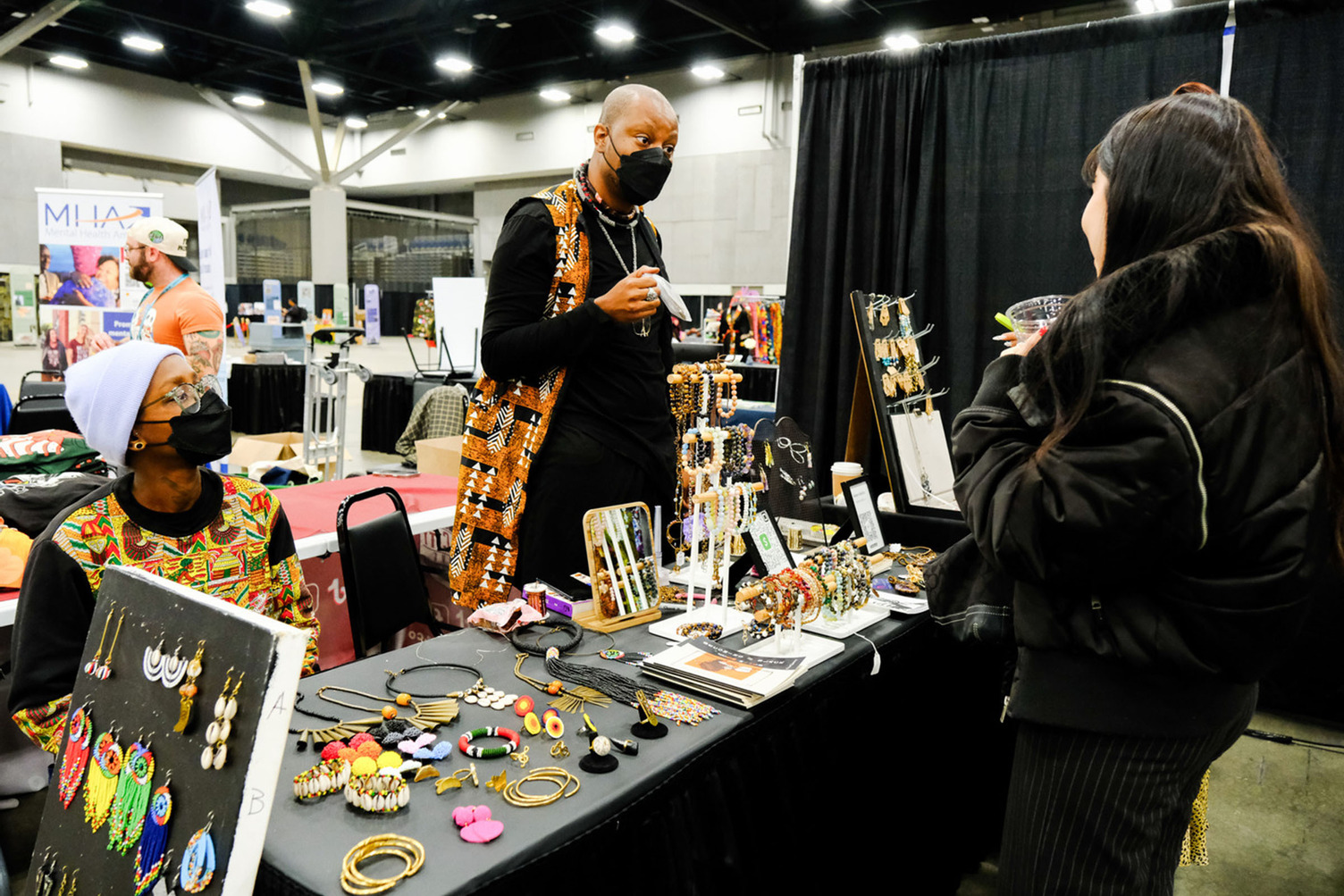 A person speaking to a vendor at table covered with colorful and beaded jewelry.