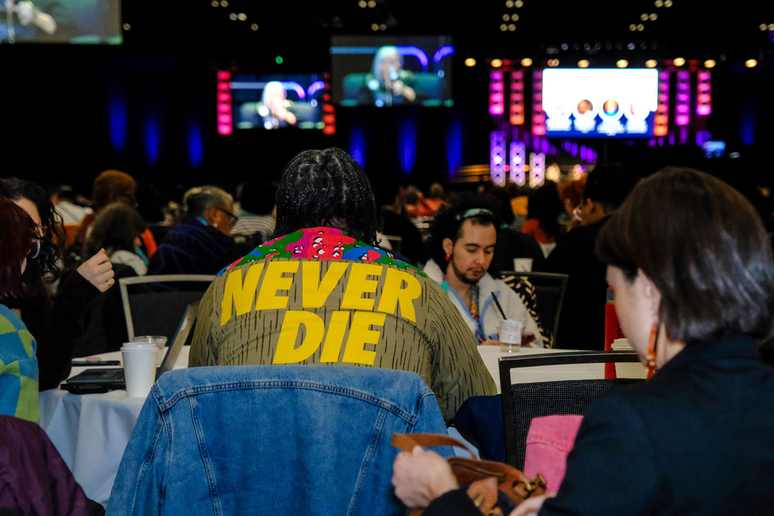 A person seated at a table with a shirt reading “Never Die.”