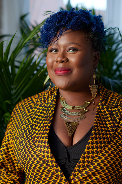 Smiling person with dark blue hair wearing a yellow and black scale-patterned jacket.