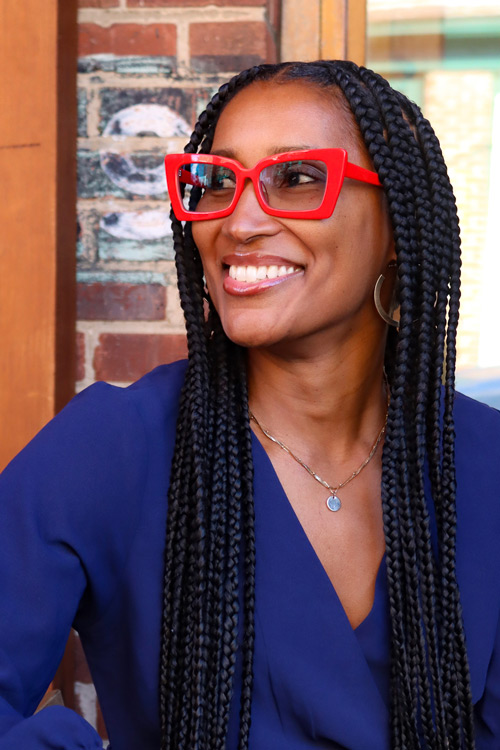 Smiling person with long dark braids and red-framed glasses.