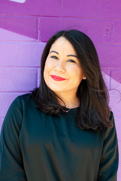 Smiling person in black shirt leaning against a two tone purple wall.