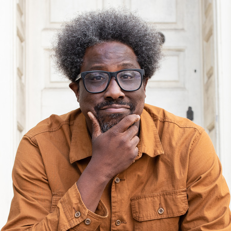 W. Kamau Bell in orang shirt with a hand on his chin.