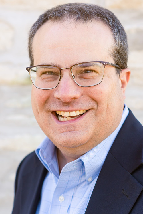 Person wearing glasses smiling in blue shirt and jacket.