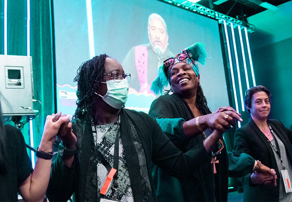 Four people holding hands in front of a stage with speaker on screen in background.