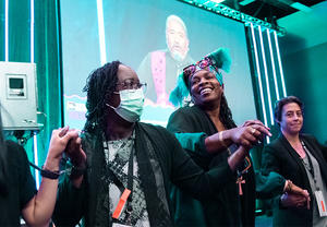 Four people holding hands in front of a stage with speaker on screen in background.