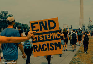 This is a photo of protestors, which includes one person holding up a sign that says. "End Systemic Racism."