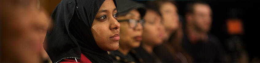 Person with head covering sitting in audience with look of interest.