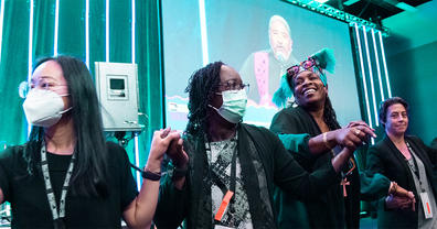 Four people holding hands in front of a stage with speaker on screen in background.