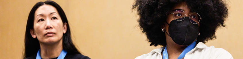 Two people looking on, one wearing black mask.